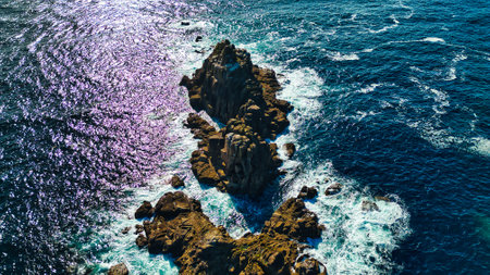 Aerial view of rocky formations surrounded by vibrant blue ocean waves. The sunlight reflects off the water, creating a shimmering effect. The rugged coastline features various textures and colors, highlighting the natural beauty of the seascape.の写真素材