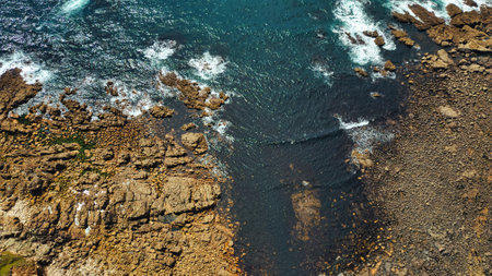 Aerial view of rocky coastline with waves crashing against the shore. The contrast between the dark water and light-colored rocks creates a striking visual. The scene captures the natural beauty of the ocean and rugged terrain.の写真素材