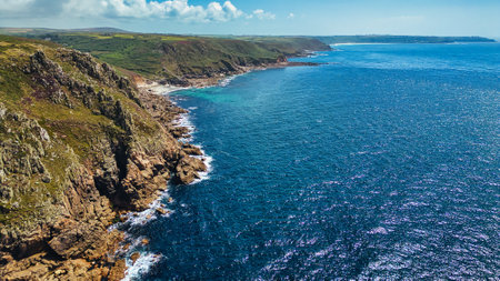 Aerial view of a rugged coastline with cliffs and clear blue waters. The landscape features green hills in the background, rocky shores, and a serene ocean. The sky is partly cloudy, enhancing the natural beauty of the scene.の写真素材