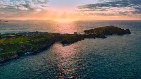 A stunning aerial view of a coastal landscape during sunset. The sun's rays illuminate the water, creating a shimmering effect. The coastline features rocky cliffs and a small settlement, with lush greenery on the land. The sky is filled with soft clouds, enhancing the tranquil atmosphere.の写真素材