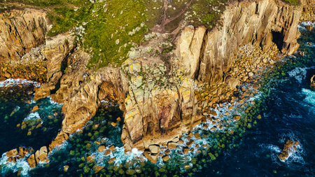 Aerial view of rugged coastal cliffs with green vegetation and rocky shoreline. The ocean waves crash against the rocks, creating a dynamic seascape. Clear blue water contrasts with the earthy tones of the cliffs and rocks.の写真素材