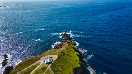 Aerial view of a coastal landscape featuring a rocky cliff extending into the ocean. The vibrant blue water contrasts with the green grass on the cliff. A small building is visible at the cliff's edge, surrounded by scenic views of the sea and distant landforms.の写真素材