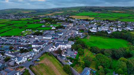 Aerial view of a small village surrounded by lush green fields. The village features a mix of residential and commercial buildings, with a parking area visible. The landscape includes rolling hills and farmland in the background, creating a picturesque rural scene.の写真素材