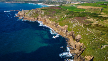 Aerial view of a rugged coastline with cliffs, clear blue waters, and green fields. The landscape features rocky outcrops and a winding path along the cliffs, with a distant view of a coastal village.の写真素材