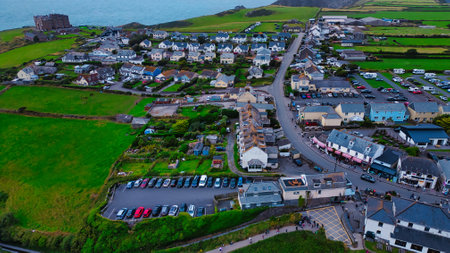 Aerial view of a coastal village with a mix of residential and commercial buildings. The landscape features green fields, parked cars, and a road leading to the sea. The ocean is visible in the background, with a few boats on the water.の写真素材