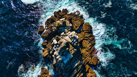 Aerial view of a rocky coastline with waves crashing against the rocks. The water is deep blue with...
