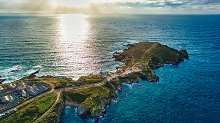 Aerial view of a coastal landscape featuring a peninsula extending into the ocean. The sun is setting, casting a golden glow over the water. Small houses are visible along the shore, with a winding path leading to the tip of the peninsula.の写真素材