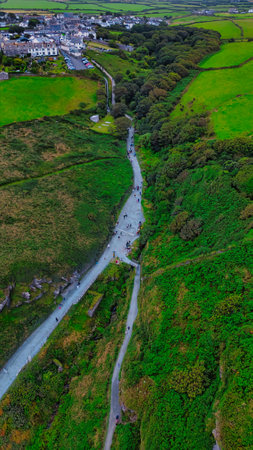 Aerial view of a lush green valley with a winding path, surrounded by trees and hills. In the background, a small town is visible, with houses and roads. People can be seen walking along the path, enjoying the scenic landscape.の写真素材