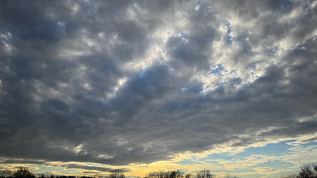 Overcast sky with predominantly dark gray and lighter gray clouds.  Patches of blue sky are visible through breaks in the cloud cover. A low horizon line shows silhouetted trees.の写真素材