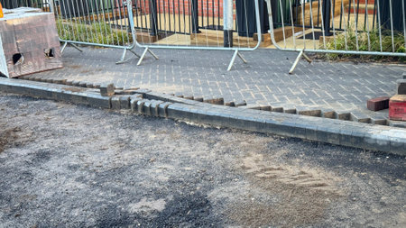 A section of newly laid pavement shows damaged kerbing and paving stones.  Loose stones and dirt are scattered nearby. Temporary metal barriers are visible in the background.の写真素材