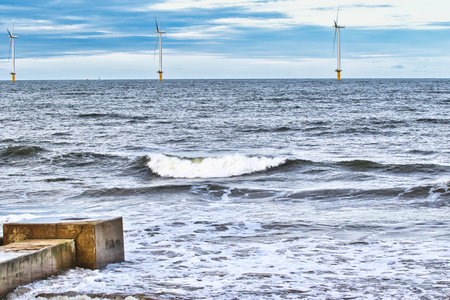 Offshore wind turbines stand in a line on a dark-blue sea.  Small waves break gently near a concrete seawall in the foreground. The sky is mostly cloudy in Redcar, UK.の写真素材