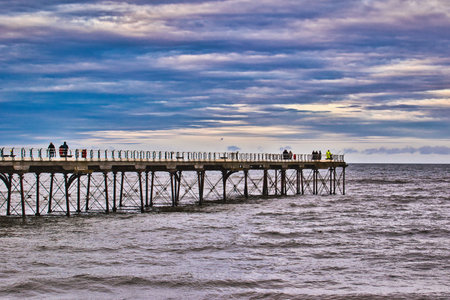 A long pier extends over a choppy sea under a cloudy sky.  Several people are visible on the pier, seemingly fishing. The overall color palette is muted blues and grays in Saltburn-by-the-Sea, UK.の写真素材