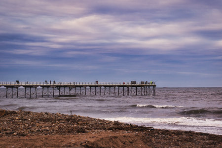 A long pier extends over a grey-blue sea under a cloudy sky.  People are visible on the pier, silhouetted against the lighter sky.  A sandy beach with debris is in the foreground.の写真素材
