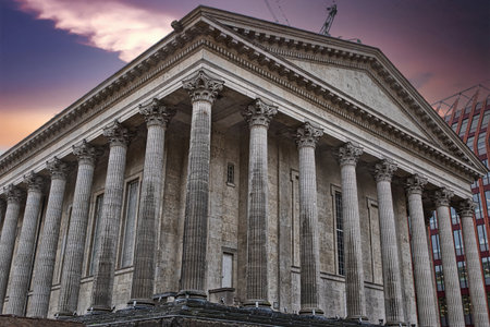 A low-angle view of a classical-style building, featuring numerous large grey columns and intricate...