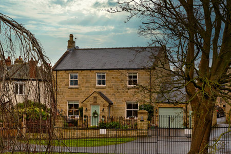 A two-story stone house with a light teal front door, flanked by windows and a detached garage.  The house is set back behind a black metal fence. A bare tree dominates the right side of the image in Knaresborough, UK.の写真素材