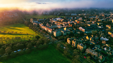Aerial view of a city neighborhood at sunset, showcasing a park, residential buildings, streets, and a light fog or mist rolling over the landscape. Warm sunset light illuminates the scene.の写真素材