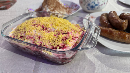 A glass dish of layered beet salad topped with shredded egg yolk sits on a table next to a plate of cooked sausages.  Other food items are visible in the background.の写真素材