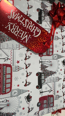 A Christmas gift wrapped in white paper featuring a London theme with red glitter accents on buses, the London Eye, and Big Ben. A red glitter gift tag says "Merry Christmas." A red bow is attached.の写真素材