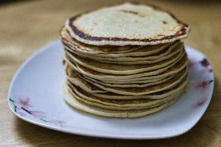 A stack of numerous light brown pancakes, showing slightly browned edges, sits on a white square plate with a floral design.  The plate rests on a light brown wooden surface.の写真素材