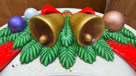 Close-up of a wet, decorative Christmas arrangement featuring two gold bells, red bows, green foliage, and multicolored ornaments.  The piece appears to be made of plastic or resin.の写真素材