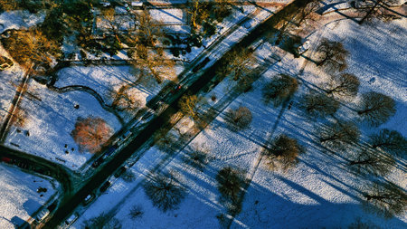 Aerial view of a snow-covered park with a road cutting diagonally through it.  Trees and their long shadows are prominent.  Several cars are visible on the road  in Harrogate, UK.の写真素材
