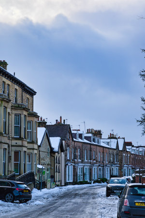 Snow-covered terraced houses line a residential street.  Two cars are parked on the snow-covered road. The sky is partly cloudy, light blue in Harrogate, UK.の写真素材