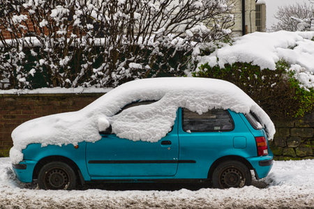 A teal-colored hatchback car is parked on a snow-covered street, heavily laden with snow on its roof and hood.  The car is situated in front of a snow-dusted brick wall and shrubs in Harrogate, UK.の写真素材