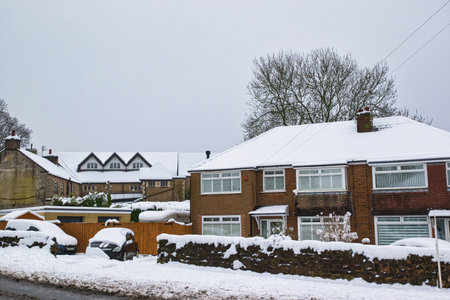 Snow-covered residential homes and cars. A two-story brick house with snow-laden roof is the focal point, situated next to a stone wall and other snow-covered buildings in Harrogate, UK.の写真素材