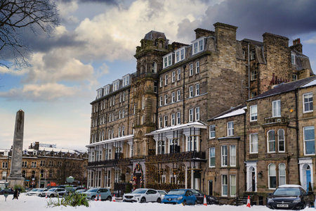 Snow-covered Harrogate town square features a large stone hotel adorned with Christmas lights, adjacent buildings, parked cars, and a tall obelisk in Harrogate, UK.の写真素材