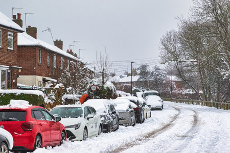 Snow-covered cars parked along a residential street lined with brick houses.  Heavy snowfall has accumulated on rooftops, vehicles, and vegetation. A person is visible clearing snow from a car in Harrogate, UK.の写真素材