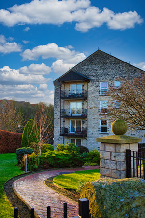 Three-story stone apartment building with balconies, landscaped grounds, and brick pathway. Sunny day with partly cloudy blue sky in Wetherby, UK.の写真素材
