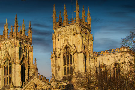 Two ornate, Gothic-style cathedral towers dominate the frame against a blue, slightly cloudy sky. Warm light illuminates the stone structures.の写真素材