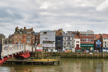 A street of buildings of varying styles and colors is lined along a waterway with a bridge in the foreground, under a cloudy sky.の写真素材