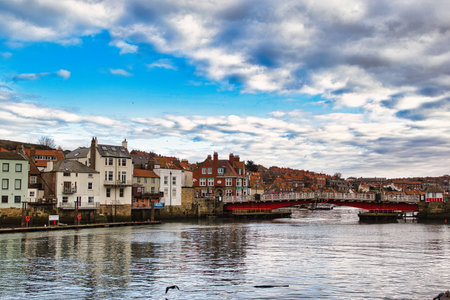 Waterfront view of town buildings along a river with a red bridge, under a partly cloudy sky reflecting in the water in Whitby, UK.の写真素材