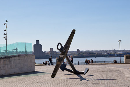 A large anchor sits on a brick plaza with a river and city skyline visible in the background. People are walking and sitting in the public space in Liverpool, UK.の写真素材