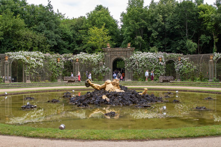 A golden statue lies partially submerged in a pond in a formal garden with trellises, arches, and lush green foliage at Palace of Versailles, Paris.の写真素材