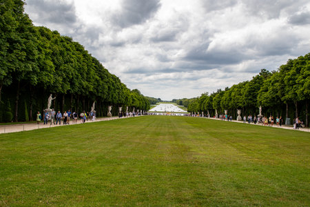 A symmetrical view of a large lawn bordered by trees, sculptures, and paths filled with people,...