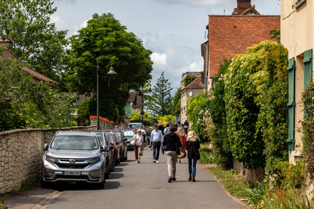 A street scene with parked cars on the left, people walking, and buildings with greenery on both sides of the street in Giverny, France.の写真素材