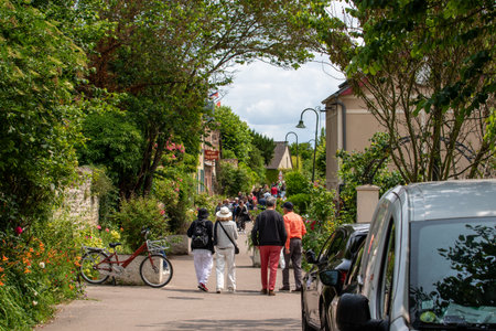 A village street view with people walking along the road, flanked by lush greenery and parked cars under a partly cloudy sky in Giverny, France.の写真素材