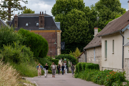 A group of people walks down a paved road lined with buildings and greenery under an overcast sky in Giverny, France.の写真素材
