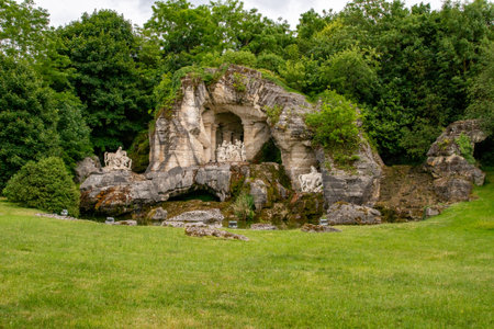 Landscape view of an artificial grotto structure made of stone, with sculptures, set within a grassy area and surrounded by trees at Palace of Versailles, Paris.の写真素材