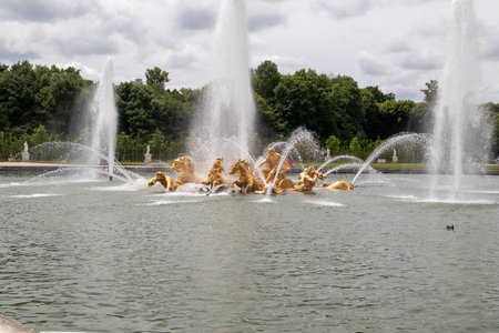 Image shows the Chariot of Apollo fountain in Versailles, featuring golden statues amidst water jets in a pond with trees in the background.の写真素材