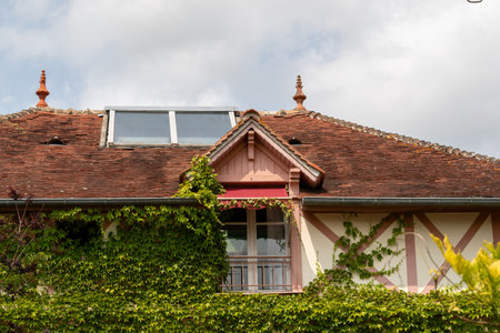 Exterior view of a building featuring a red-tiled roof, dormer window, and ivy-covered facade under a cloudy sky in Giverny, France.の写真素材