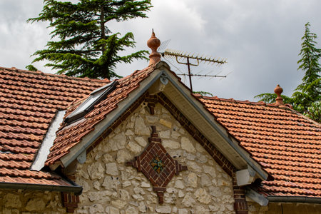 Close-up of a building with an orange tile roof, stone gable end, and television antenna against a cloudy sky. Trees are visible in the background in Giverny, France.の写真素材