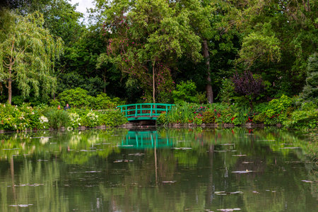 Lush green foliage frames a reflective pond with a bright green arched footbridge in the background in Giverny, France.の写真素材