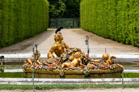 Gilded statue of figures on a floral raft in a pool, with a black bird perched on one figure's head. Hedged path in the background at Versailles Gardens, Paris.の写真素材