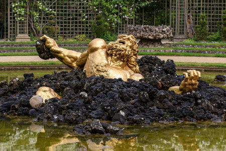 Gilded statue of struggling figure emerging from black rocks, located in a pool within a garden setting at Versailles Gardens, Paris.の写真素材