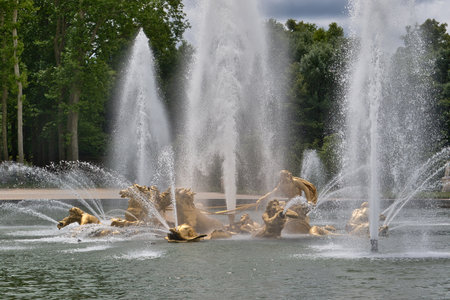 Golden sculptures in a fountain with tall water jets. Trees are in the background under a cloudy sky at Versailles Gardens, Paris.の写真素材