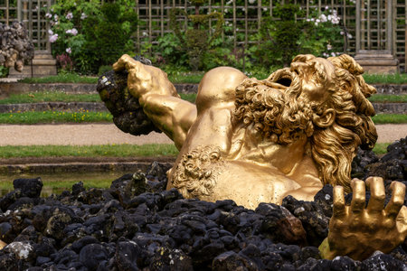 Golden statue of anguished figure atop black rocks, grasping a stone, in an outdoor garden setting at Versailles Gardens, Paris.の写真素材