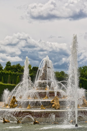 A tiered fountain with golden statues gushes water upwards under a cloudy sky, framed by trees and hedges at Versailles Gardens, Paris.の写真素材